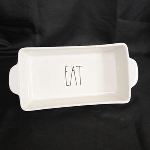 May include: A rectangular white ceramic serving dish with rounded handles. The word "EAT" is printed in black, centered on the inside of the dish. The dish is on a black surface.