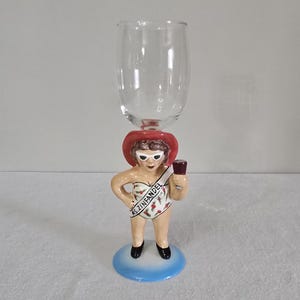 May include: A clear glass wine glass with a ceramic figurine of a woman wearing a red hat, white sunglasses, and a swimsuit with the text "Ms. Zinfandel".