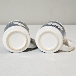 May include: Two white ceramic mugs positioned upside down, revealing their circular bases. The exterior of each mug features a dark blue band decorated with a repeating white floral pattern and a white silhouette of a dog in a running pose.
