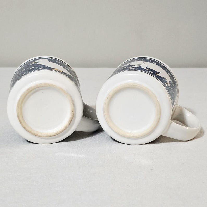May include: Two white ceramic mugs positioned upside down, revealing their circular bases. The exterior of each mug features a dark blue band decorated with a repeating white floral pattern and a white silhouette of a dog in a running pose.