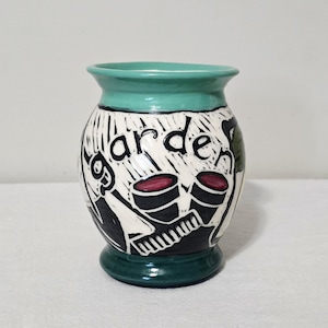 May include: A ceramic vase with a flared rim and base in teal. The body features a white background with black carved illustrations of gardening tools and the word garden in black script. Two red circular accents are visible on the tools.