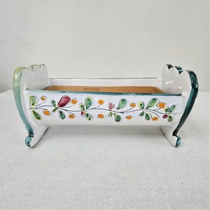 Peut inclure: Une jardinière rectangulaire en céramique à finition émaillée blanche, ornée d'un motif de vigne peint à la main avec des feuilles vertes et des baies orange sur le devant. Elle possède des supports latéraux incurvés avec des bords verts.
