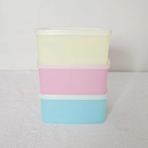 May include: Three rectangular food storage containers stacked on top of each other. The top container is yellow, the middle one is pink, and the bottom one is blue. Each container has a white lid.