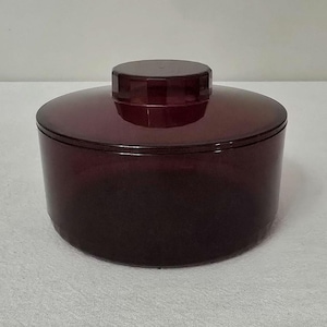 May include: A round, dark burgundy container with a matching lid. The container has a cylindrical base and a slightly domed lid. The lid sits flush on top of the container. The container is made of a translucent material.