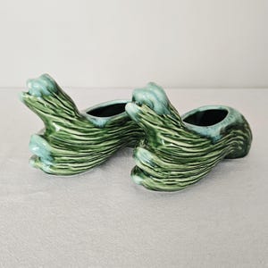 May include: Two ceramic planters with an unusual, organic form. The planters are a mix of green and teal, featuring a textured, wave-like design. The inside of each planter is a dark teal, providing a strong contrast. These decorative planters are perfect for small plants.