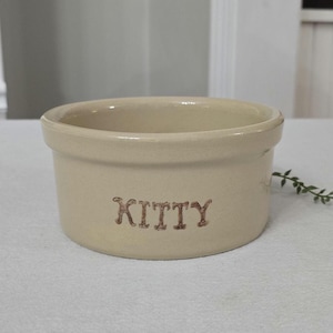 May include: A cream-coloured ceramic pet bowl with the word "KITTY" in brown lettering. The bowl has a rounded shape and a slightly flared rim. A small sprig of greenery is visible in the background.