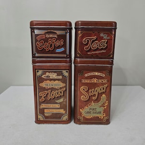 May include: Four rectangular brown metal storage tins with lids. The tins are labeled "Coffee", "Tea", "Flour", and "Sugar" in decorative fonts. The flour tin reads "Golden Harvest" and the sugar tin reads "Crystal Cane".