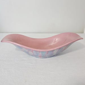 May include: A pink ceramic bowl with a wavy, elongated shape. The interior is a solid, soft pink, while the exterior rim features a blend of pastel colors, including pink, blue, and white. The bowl is designed for decorative or serving purposes.