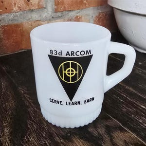May include: A white milk glass mug featuring a black triangle graphic with a yellow circular emblem inside. The text 83d ARCOM is printed above the triangle, and the words SERVE, LEARN, EARN are printed below. The mug has a ribbed base and handle.