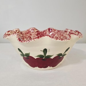 May include: Cream-colored ceramic bowl with a ruffled edge and apple design. The rim has a red speckled pattern. The bowl features three red apples with green leaves painted on the front. A decorative kitchenware item.