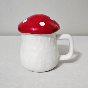 May include: A ceramic mug shaped like a mushroom with a white base and a glossy red lid featuring white spots. The mug has a rounded body with a textured surface and a sturdy handle on the right side, designed to hold hot or cold beverages.