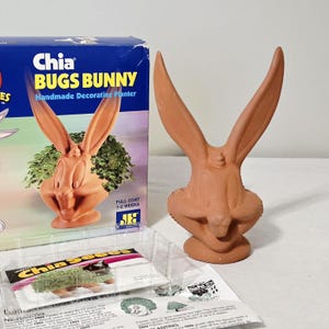 May include: A terracotta Bugs Bunny planter featuring long ears and a sculpted face. The product is accompanied by its original blue cardboard packaging labeled Chia Bugs Bunny Handmade Decorative Planter, a plastic tray, and instructions.