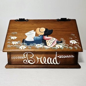 May include: A wooden bread box with a slanted lid featuring a hand-painted illustration of two figures sitting in a field of white daisies. One figure holds a newspaper labelled THE BUDGET. The word Bread is painted in white script on the front.