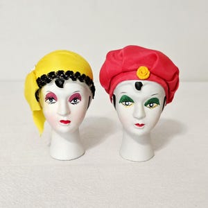 May include: Two ceramic head busts featuring painted facial features. The left bust wears a yellow fabric head wrap with a black trim. The right bust wears a red fabric beret-style hat adorned with a small yellow fabric rose centered on the front.