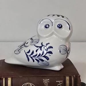 May include: A white ceramic owl figurine with blue floral designs. The owl is perched on a brown book. The owl has large, round eyes and a detailed feather pattern. A decorative item.