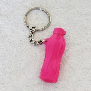 May include: A bright pink keychain featuring a miniature plastic water bottle charm attached to a silver-toned metal split ring by a short chain. The bottle charm has a textured, contoured surface and a small cap at the top, reflecting light.