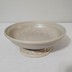 May include: A light beige ceramic pedestal bowl with a speckled finish. The interior base features an embossed vine and leaf pattern. The circular base of the pedestal is decorated with a ring of small, raised circular dots around the edge.