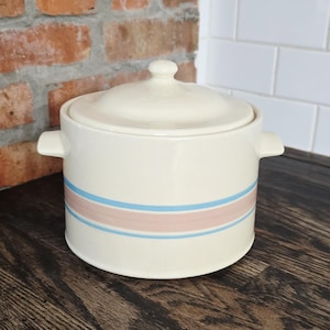 May include: Cream-coloured ceramic casserole dish with a lid and two handles. The dish is decorated with two horizontal stripes, one light blue and one pink. The dish is on a dark wooden surface.