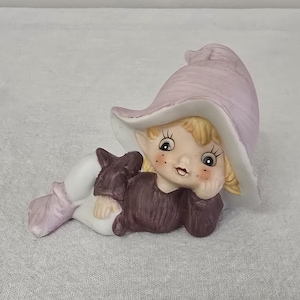 May include: A porcelain figurine of a girl elf wearing a purple hat and a brown dress. She is lying on her side with her head resting on her hand.