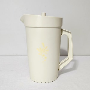 May include: A cream-coloured plastic jug with a matching lid, a side handle, and a pouring spout. The front of the jug features a subtle, light yellow floral graphic design consisting of small leaves and dots arranged in a vertical pattern.