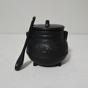May include: A black ceramic cauldron with a lid and a matching spoon. The cauldron has three small feet and two handles. The words "Harry Potter" are embossed on the front. The spoon is resting against the cauldron.