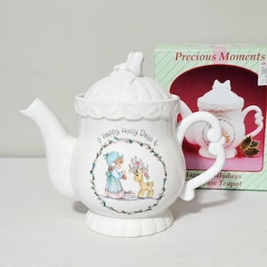 May include: A white ceramic teapot with a lid, featuring a decorative illustration of a child and a reindeer within a holly wreath. The text Happy Holly Days is printed above the illustration. The teapot has a curved handle and spout.
