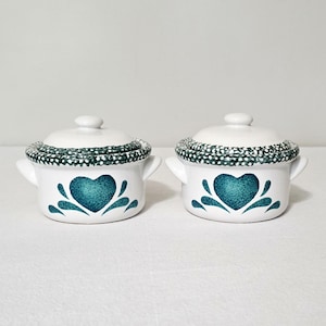 May include: Two white ceramic lidded pots with side handles, each featuring a dark teal heart-shaped design on the front and a dark teal rim with white polka dots. The lids are white with a small knob handle on top, matching the pots' design.