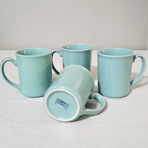 May include: Four turquoise ceramic mugs with curved handles are arranged on a white surface. One mug is overturned, revealing the Corning brand stamp on the base. The mugs have a simple, classic design.