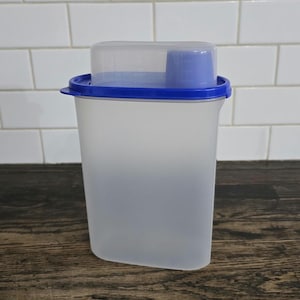 May include: A clear, rectangular food storage container with a blue lid and a blue measuring cup. The container is made of translucent plastic and sits on a dark wooden surface. The lid is clear with a blue rim.