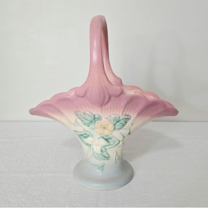 May include: A ceramic basket with a tall, arched handle and a flared, scalloped rim. The piece features a gradient color scheme transitioning from a soft pink at the top to a cream and pale blue base, decorated with embossed floral motifs.