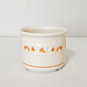 May include: A cream-colored ceramic crock featuring a repeating pattern of orange, yellow, and white candy corn motifs around the center. The cylindrical container has a smooth finish and a single thin orange horizontal line near the base.
