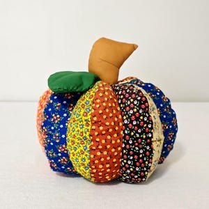 May include: A stuffed fabric pumpkin decoration made from vertical segments of various floral patterned cotton fabrics in blue, yellow, orange, black, and cream colors, featuring a solid brown fabric stem and a single green fabric leaf.