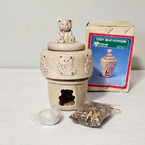 May include: A beige ceramic potpourri burner featuring a teddy bear figurine on the lid and embossed teddy bear motifs around the rim. The base has a bear-shaped cutout. Includes a small tea light candle, a bag of potpourri, and the original box.