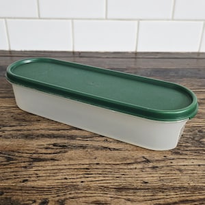 May include: A rectangular, translucent plastic food storage container with a dark green lid. The container is empty and sits on a wooden surface. The lid is oval-shaped and fits snugly on the container.
