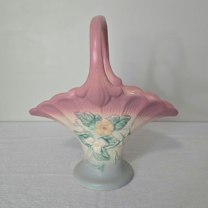 May include: A vintage ceramic basket with a handle, in pink and white. The basket has a flared, petal-like design and is decorated with a floral motif in yellow, green, and white. The base is a soft blue. A decorative item.