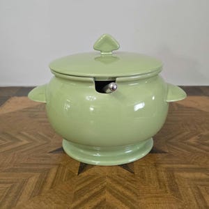 May include: A pale green ceramic soup tureen with a lid and a ladle holder. The tureen has a rounded body and a flared base. The lid has a small knob in the shape of a diamond.