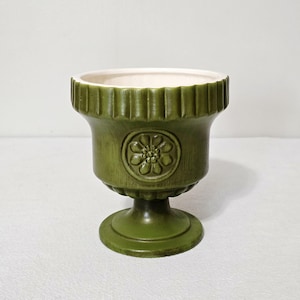 May include: An olive green ceramic pedestal planter featuring a fluted rim and a circular medallion with a raised floral design on the front. The interior of the vessel is finished in a smooth white glaze, contrasting with the textured green exterior.