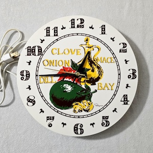 May include: A round white wall clock with black numerals and a white power cord. The face features a central illustration of a yellow oil cruet, a green avocado, and red peppers, with the yellow text CLOVE, ONION, MACE, DILL, and BAY printed.