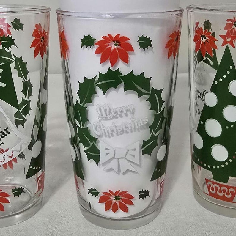 May include: Three clear glass tumblers with Christmas-themed designs. Each glass features red poinsettias, green holly, and the words "Merry Christmas" in a wreath. One glass has a Christmas tree design.