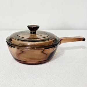 Peut inclure: Une casserole en verre ambré translucide avec un couvercle assorti. Ce récipient de cuisson rond présente une longue poignée unique s'étendant sur le côté et un bouton circulaire au centre du couvercle, avec une finition lisse et polie.