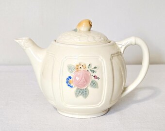 May include: A cream-coloured ceramic teapot featuring a raised floral motif on the front with a pink rose, blue petals, and green leaves. The lid is topped with a small, rounded finial, and the body has a panelled design with a curved handle and spout.
