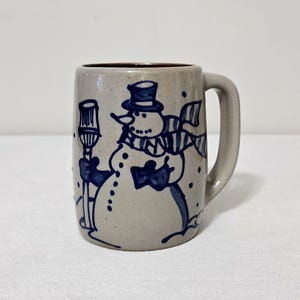 May include: A beige ceramic mug featuring a hand-painted dark blue illustration of a snowman wearing a top hat and a striped scarf. The snowman holds a broom, and the design includes small dots representing falling snow on the speckled surface.