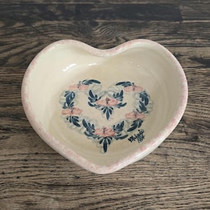 May include: Heart-shaped ceramic dish with a cream-colored interior and a pink rim. The dish features a floral design in the center, with pink and blue flowers arranged in a heart shape. The bottom right corner has a signature and the year 1994.