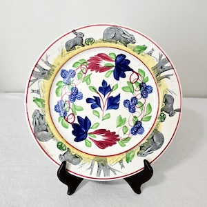 May include: A decorative ceramic plate featuring a whimsical design. The plate has a white background with a red and yellow border. It is adorned with illustrations of rabbits, flowers, and foliage. The plate is displayed on a black stand.