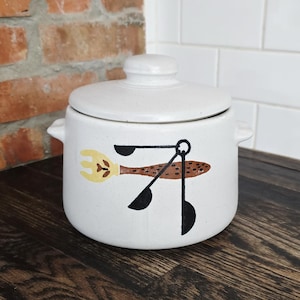 May include: A white ceramic kitchen canister with a lid. The canister features a whimsical design of a fork, spoon, and measuring spoons in brown, yellow, and black. The canister has a speckled finish and small side handles.