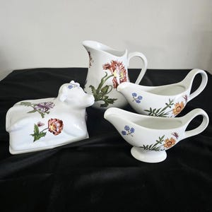 May include: A white ceramic cow-shaped butter dish, a tall pitcher, and two gravy boats, all decorated with colorful botanical floral patterns including red poppies, orange daisies, and small blue flowers, arranged together on a dark surface.