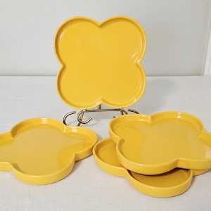 May include: A collection of bright yellow, clover-shaped ceramic plates. One plate is displayed on a gold-colored metal stand, while several others are arranged in front. The plates have a glossy finish and a slightly raised edge.