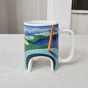 May include: White ceramic mug with a golf course design. The mug features a golf course scene with a flag, water, and an orange pole. The handle is a square shape, and the base has a unique curved design.