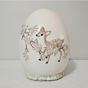 May include: A large, egg-shaped ornament with a white surface. It features a painted design of a fawn, a blue bird, and pink flowers on branches. The base is a textured, light green colour.
