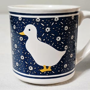 May include: A white ceramic mug featuring a dark blue background decorated with a repeating pattern of small white flowers. A large, solid white duck with a yellow beak and yellow feet is centered on the front of the mug, which has a handle.